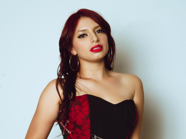 LeticiaFlorenz with red lipstick, hoop earrings, and a strapless red and black lace dress, showing her tits and a slight smirk. She has red hair and a sexy look.