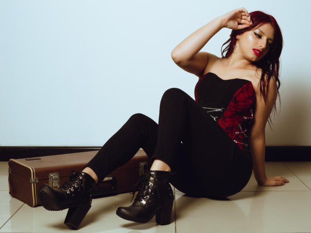 LeticiaFlorenz, avec des cheveux roux, un haut noir sans bretelles, un corset en dentelle rouge, un pantalon noir et des bottes noires à lacets, assise sur un sol carrelé, une main dans les cheveux, regardant vers le bas..