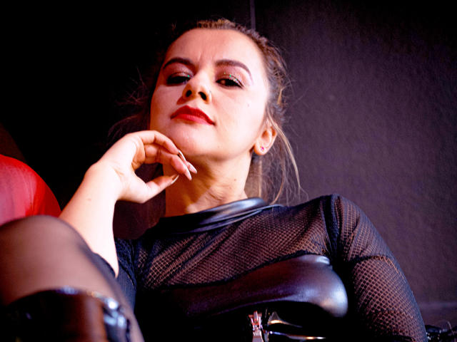 PerlaDivaio with red lipstick, dark mesh top, black leather, and a smug expression, hand near chin, looking down at the camera. Her tits are slightly visible through the mesh.