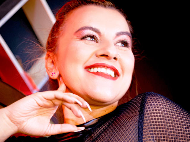 PerlaDivaio's smiling face with red lipstick, pink earrings, and white nails, wearing a black mesh top, looking up with her hand near her chin, red and black background.
