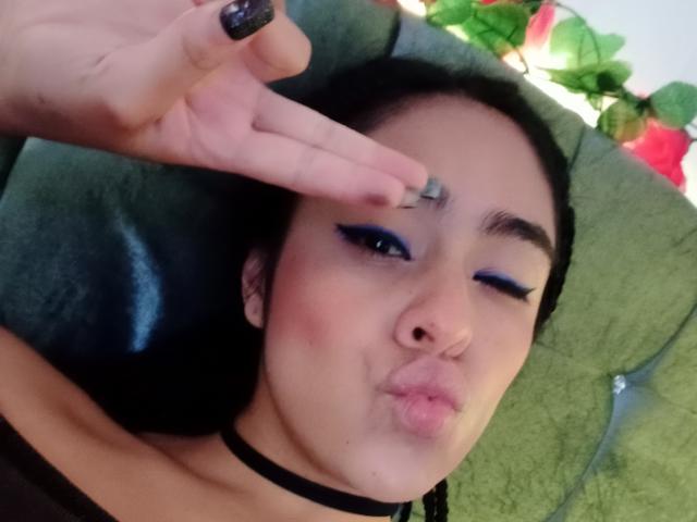 StellaBeckker with black choker, blue eyeliner, pouty lips, and black nail polish, making a peace sign near her eyebrow, lying on a green cushion.