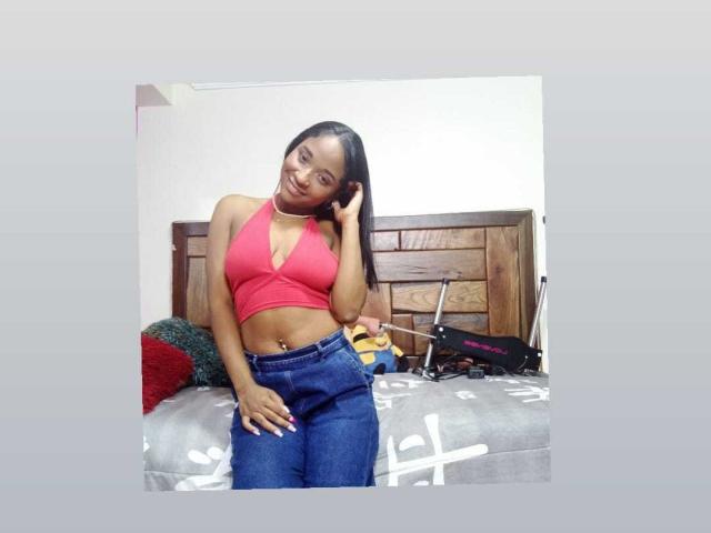 MelanyScoth with small tits, wearing a red halter top and blue jeans, standing on a bed with a wooden headboard, smiling, hand on her head, black hair.