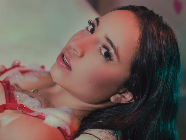 GiannaRusell, con el cabello negro mojado, un sujetador rojo y los pezones cubiertos de jabón, mirando seductoramente a la cámara, con los labios ligeramente entreabiertos, ojos azules, y una luz rosada en su rostro..