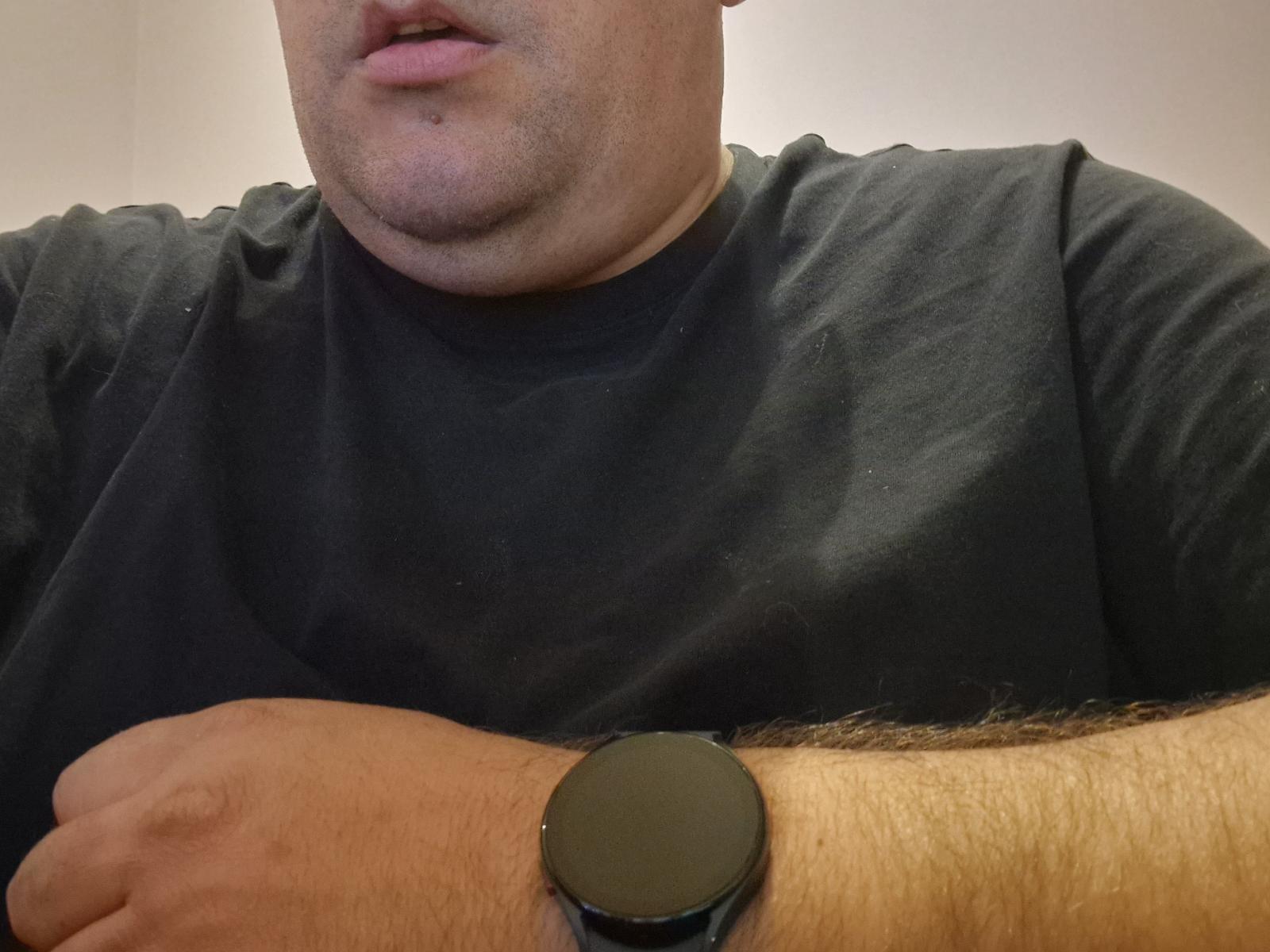 FreddyJames, un homme corpulent portant une chemise noire et une montre intelligente noire à son poignet poilu, a les lèvres légèrement entrouvertes, laissant entrevoir un peu de sa lèvre supérieure..