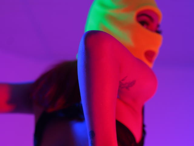 LeahMarie with colorful neon lights, wearing a mask, exposing her tits, in black lingerie, bent over, showcasing her ass and tattoo on her arm.
