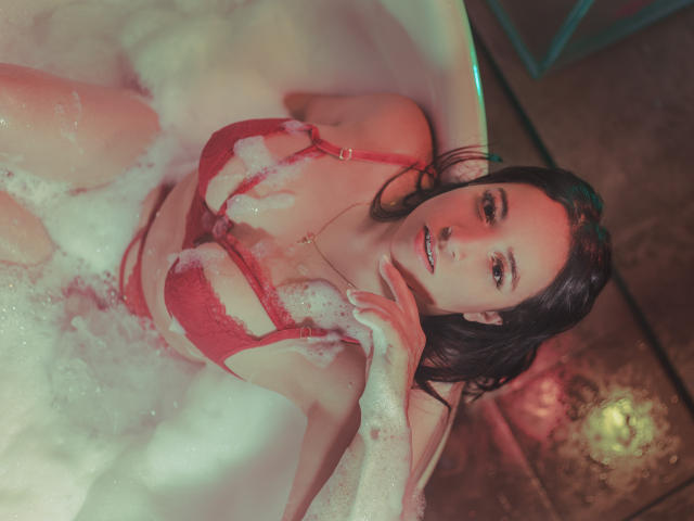 GiannaRusell, con el cabello oscuro y mojado, lencería roja y burbujas de jabón en una bañera, con la mano cerca de la boca, mirando hacia arriba, con reflejos de luz rosa y verde. Sus pechos son visibles..