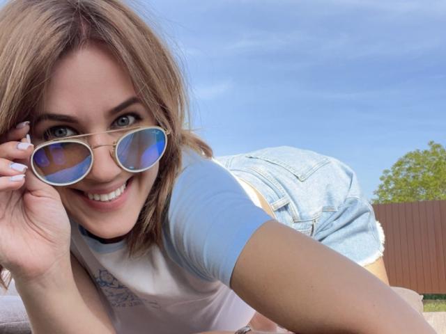 Misstyk with blue-tinted aviator sunglasses, smiling, wearing a light blue shirt and denim shorts, leaning forward, showing her ass. Her brown hair and green eyes are visible.