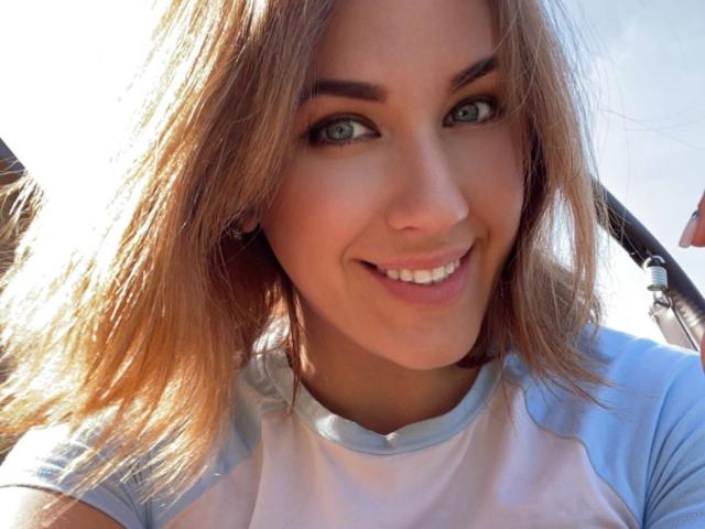 Misstyk has blonde hair, blue eyes, and a cute smile. She's wearing a light blue and white raglan shirt. Her tits are small and her pussy is not visible.