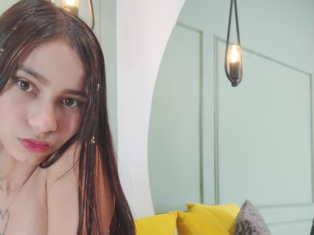 CarolCastroI mit langen braunen Haaren, rosafarbenem Lippenstift und dezentem Make-up, die in die Kamera lächelt, gelbe Kissen im Hintergrund, eine Lampe hängt darüber..