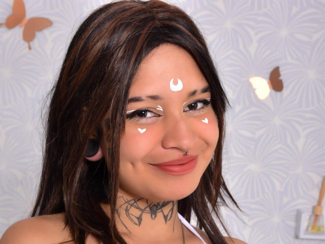 SayuriRose, com cabelo castanho, maquiagem branca no rosto, piercing no nariz, protetores de ouvido pretos e tatuagem no pescoço, sorrindo, mostrando os seios, pequenos mamilos rosados, fofa, com um fundo branco estampado..