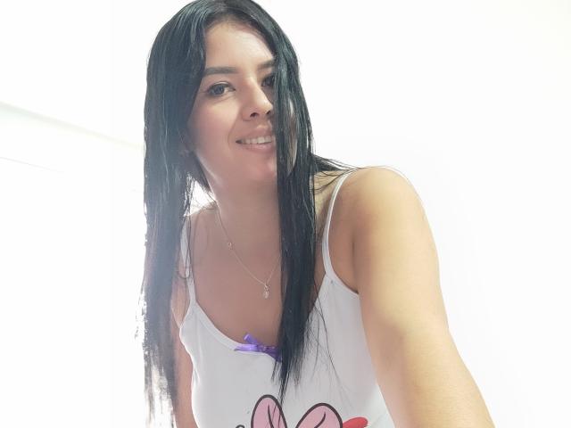 AshleyKatelyn, com longos cabelos pretos e molhados, blusa de alças branca, laço roxo, sorrindo, e uma estrutura esguia, mostrando seios pequenos e um rosto fofo, usando um colar delicado..
