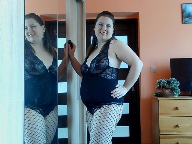 WendyStacy with brown hair, wearing black lace lingerie and fishnet stockings, standing in front of a mirror, smiling, showing her tits and ass, with one hand on the mirror.