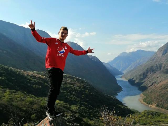 ShatSex steht auf einem Felsen, die Arme ausgestreckt, die Mittelfinger nach oben gerichtet, und trägt ein rotes Pepsi-T-Shirt, schwarze Hosen und Turnschuhe, in einem bergigen Tal mit einem Fluss..