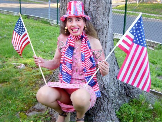ElenahHottie está agachada perto de uma árvore, segurando duas bandeiras americanas, usando um chapéu, cachecol e vestido nas cores vermelho, branco e azul, com um sorriso patriótico e brincos coloridos..