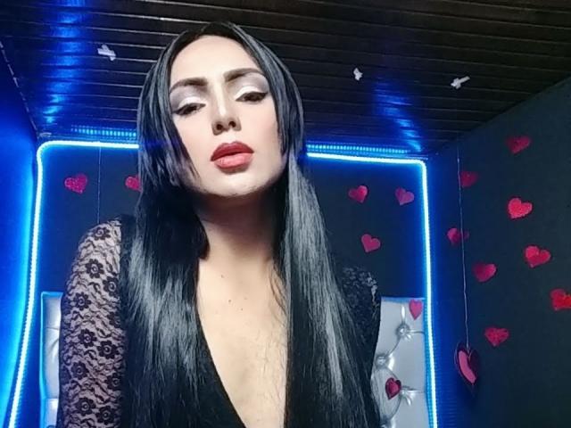 Iris Durand mit langem, schwarzem Haar, die ein schwarzes Spitzenoberteil, roten Lippenstift und einen tiefen Ausschnitt trägt, blickt nach unten, im Hintergrund blaue LED-Leuchten, rote Herzaufkleber..