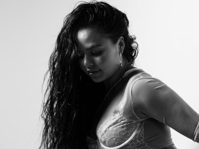 ArielleWilde with wet, dark hair, wearing a sheer, lace bra, looking down with a slight smile, large hoop earrings, gray-black monochrome, soft lighting, upper body shot.