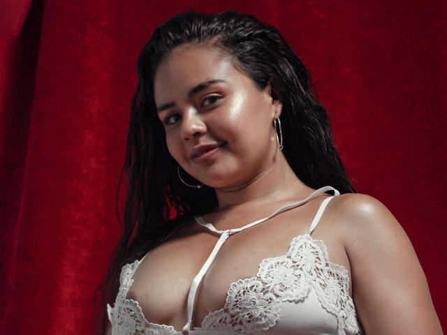 ArielleWilde with shiny skin, wet dark hair, wearing a white lace bra, showing her large tits, smiling, red background, hoop earrings.