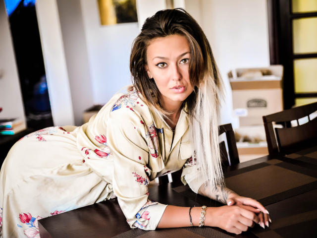 SpicyAlicia, con su largo cabello rubio oscuro, se apoya en una mesa de madera oscura, vestida con un vestido floral de color crema, y sus ojos miran fijamente a la cámara, luciendo atractiva..
