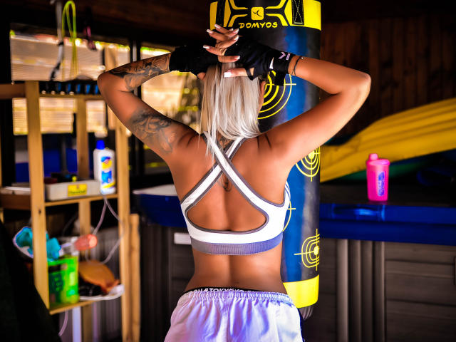 SpicyAlicia with blonde hair, back to camera, wearing a white sports bra and purple shorts, tattoo on left arm, black fingerless gloves, standing in a gym with a yellow bag.