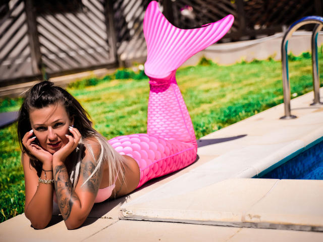 SpicyAlicia, a pink mermaid with a shiny pink tail, lays by the pool, hands on chin, smiling. She's got tattoos on her arm and blonde hair.