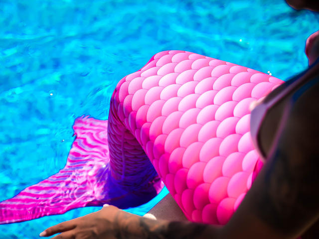 La coda rosa e lucida di SpicyAlicia, a forma di sirena e con squame tonde, è parzialmente immersa in acqua blu brillante; la sua mano è appoggiata sul bordo, mostrando un braccio con tatuaggi..