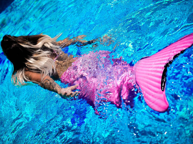 SpicyAlicia with long blonde hair, tattoos, and pink mermaid tail, floating in blue pool, arms out, tail fin visible, water rippling, sunny day, vibrant colors.