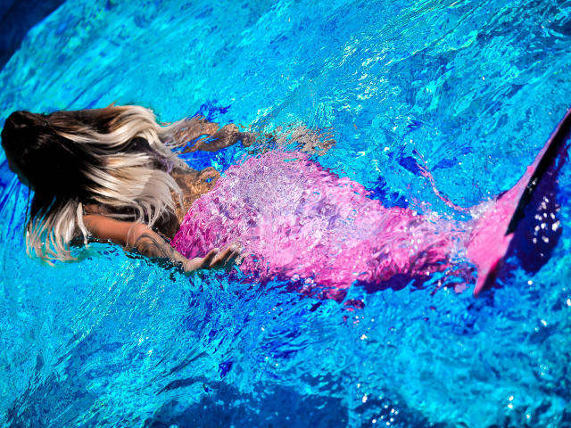 SpicyAlicia mit einem pinken Meerjungfrauen-Schwanz, die in leuchtend blauem Wasser schwebt, ihr langes blondes Haar weht im Wind, ein Tattoo auf ihrem Arm, mit dem Rücken zur Kamera, das Wasser plätschert um sie herum..