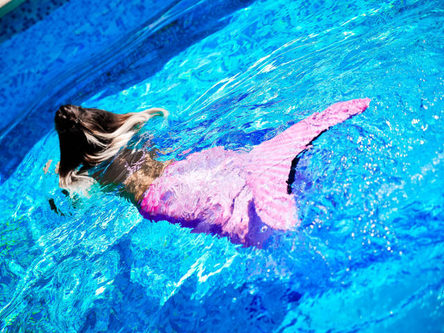 SpicyAlicia in a pool, floating face-down, pink swim bottom, brown hair, blue water, sun shining, translucent, water rippling, pink swim bottom clearly visible.