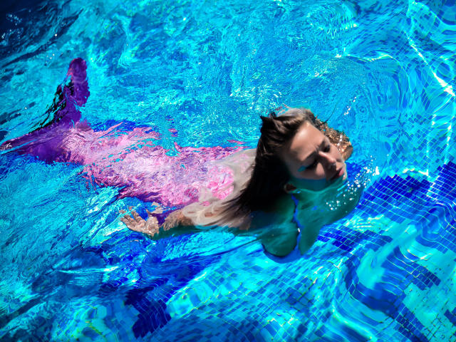 El trasero rosado de Alicia, vestido con un bikini, flota en una piscina con azulejos azules, su cabello castaño y sus pechos parcialmente sumergidos, mirando hacia arriba con una expresión relajada..