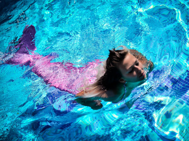 SpicyAlicia with pink mermaid tail, floating in blue pool, water rippling, eyes closed, brown hair, slightly visible boobs, purple tail fin, sunlit, underwater.