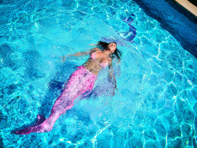 SpicyAlicia with pink mermaid tail, floating in bright blue pool, water rippling over her topless body, showing her tits, arms and legs relaxed, sun shining on her face.