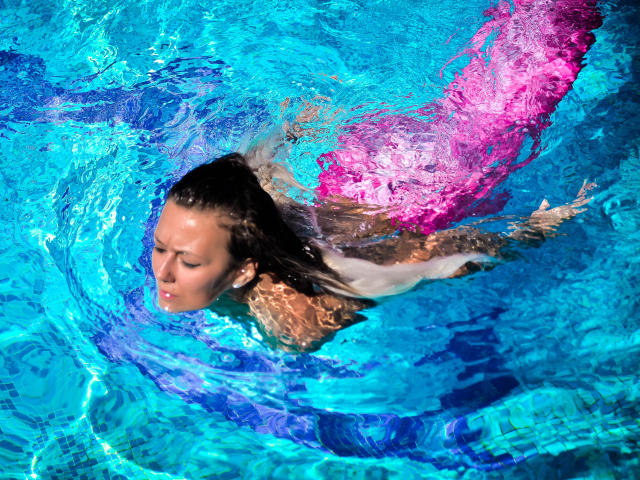 SpicyAlicia with her ass down, wearing a pink swim cap, swimming in bright blue water with her eyes closed and wet brown hair. Her tits are partially visible.