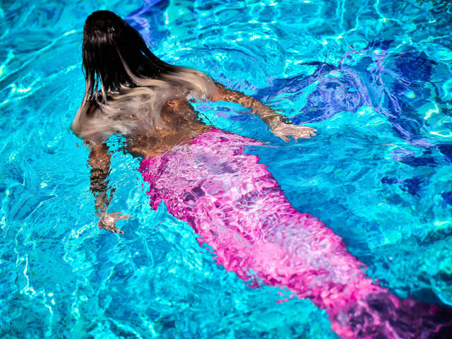 SpicyAlicia with wet, dark brown hair, back to the camera, in a bright blue pool, wearing a pink mermaid tail, arms out, water rippling around her.