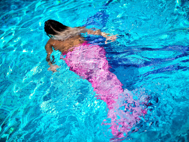 SpicyAlicias Hintern ist in einem hellrosa Badeanzug über dem blauen Wasser sichtbar, mit dem Rücken zur Kamera und dunklem, braunem Haar, während sie schwimmt..