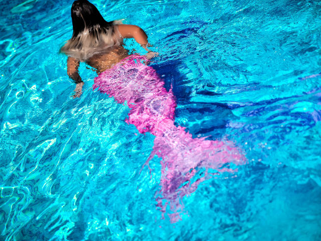 SpicyAlicia with a pink mermaid tail, her back to the camera, swimming in bright blue water, her dark hair wet, tail's pink texture contrasting with the water.
