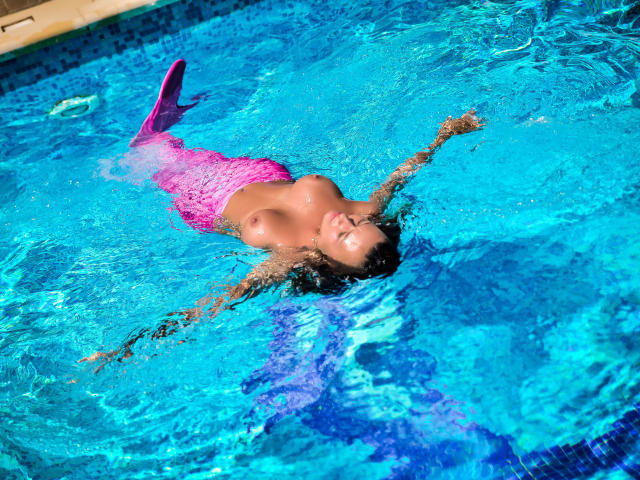 SpicyAlicia avec une queue de sirène rose, flottant dans une piscine bleue, les bras ouverts, nue, montrant ses grandes seins et ses cheveux bruns mouillés, les yeux fermés, sous une lumière du soleil vive..