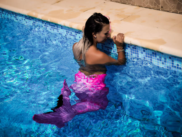 SpicyAlicia, with wet black and blonde hair, leans on a pool edge, her pink mermaid tail, blue water, and tattoos visible. Her ass is above the water.