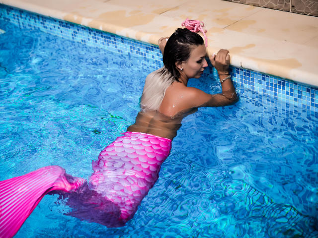 SpicyAlicia, une sirène avec une queue écailleuse rose, est appuyée contre le bord de la piscine, l'eau scintillante sur sa peau bronzée, ses cheveux blonds attachés avec un élastique rose, piscine avec des carreaux bleus, détendue, sexy..