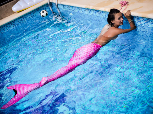SpicyAlicia with a pink mermaid tail, ass raised, floating in a blue pool, leaning on the edge, ball in water, wet hair, looking back, arms on pool edge.