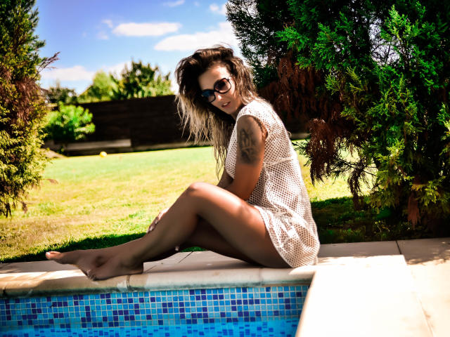 SpicyAlicia sitzt am Pool, trägt ein durchsichtiges weißes Kleid, das ihren Hintern und ihren tätowierten Arm zeigt, trägt eine Sonnenbrille und hat blondes Haar zu einem unordentlichen Dutt gebunden..