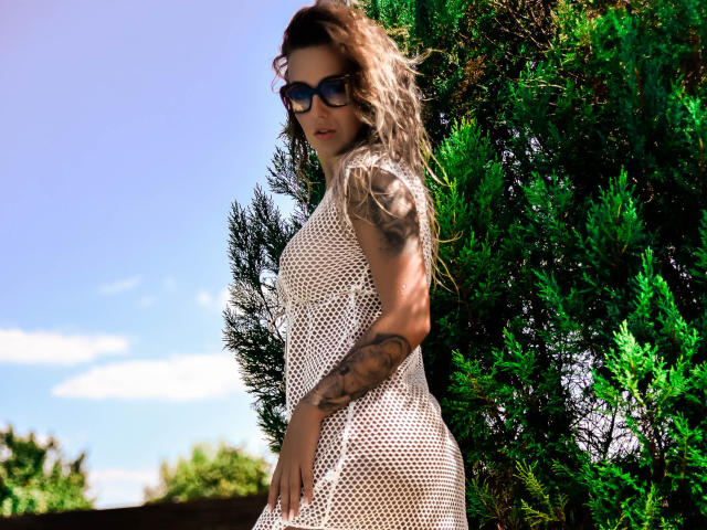 SpicyAlicia mit welligem, blondem Haar, die ein weißes Netzkleid, eine dunkle Sonnenbrille und Tattoos am rechten Arm trägt, steht neben grünen Bäumen und zeigt ihr kurviges Hinterteil..
