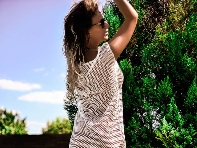 SpicyAlicia with wavy blonde hair, wearing a white mesh top, raising her arm, standing near a green bush, wearing sunglasses, sunny outdoor setting.