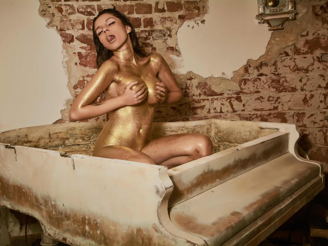 GoddessKaterina with golden body paint, sitting in a dirty white bathtub, covering her tits with one hand, head back, eyes closed, looking sexy and seductive.