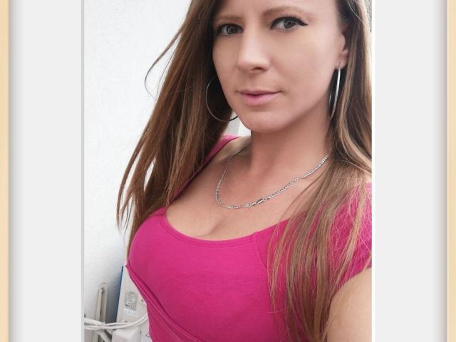 LaMadrina with brown hair, pink top, showing her tits, wearing a silver chain necklace, and large hoop earrings, has a slight smile and is looking at the camera.