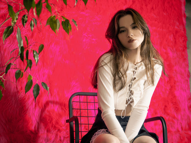Kaitlyn : Une jeune femme aux longs cheveux bruns, portant un haut blanc à lacets et une jupe noire, assise sur une chaise noire, avec un fond rose et duveteux. Une plante verte est visible sur la gauche. Elle regarde l'appareil photo avec une légère inclinaison de la tête..