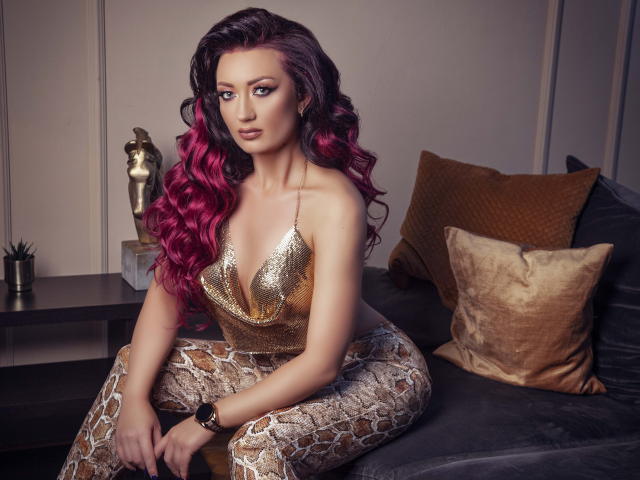 MiaBlackXO with pink and black curly hair, gold sequined top, and snake-pattern pants, sitting on a gray couch, looking at the camera with a confident expression.