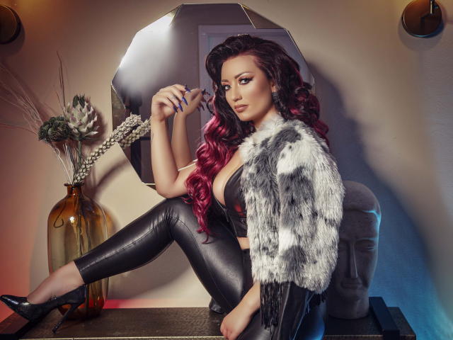 MiaBlackXO with red curls, wearing black leather pants, fur jacket, and high heels, holding a mirror, showing her tits, sitting on a table, with a vase in the background.