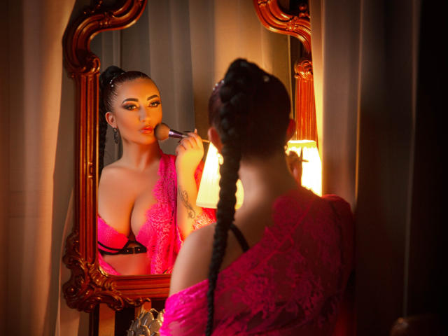 MiaBlackXO with braided hair, wearing pink lace, applying makeup in front of an ornate mirror, focusing on her tits in a pink bra, warm lamplight.