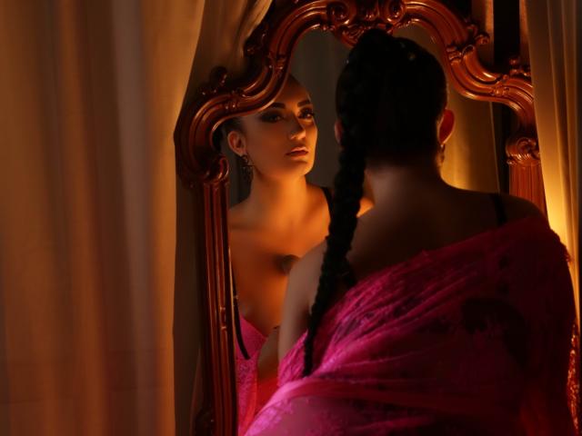 MiaBlackXO with braids, pink off-shoulder dress, reflects in ornate mirror, looking at her tits, warm light, curtains, dark brown hair, earrings, sexy, elegant.