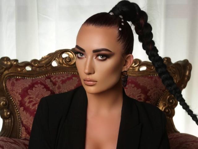 MiaBlackXO with a high ponytail, braided and beaded, wearing a black blazer, has tan skin, brown eyes, and a serious expression, sitting on an ornate red and gold chair.